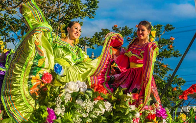 Dos bailarinas con vestidos tradicionales verde y rosa, flores en el pelo y faldas levantadas al bailar al aire libre