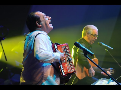 Músico tocando el acordeón y cantando en un escenario iluminado, acompañado por un guitarrista de fondo